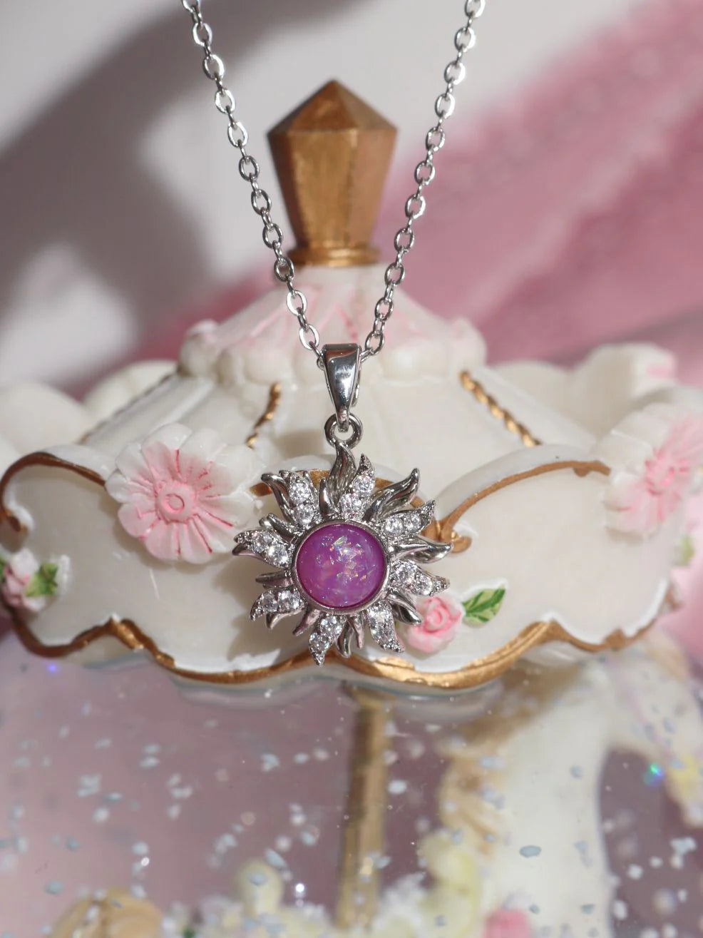 A silver-toned sunburst pendant necklace with a central oval purple gemstone and small clear stones, displayed on a decorative white and pink stand.