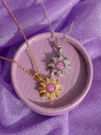 Two sunburst pendant necklaces with pink cabochon stones, one in gold and one in silver, displayed on a round lavender tray over purple fabric.