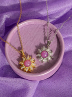 Two sunburst pendant necklaces displayed on a round lavender tray. One pendant is gold-toned with a pink cabochon stone and small clear stones around it, and the other is silver-toned with a matching pink stone and clear stones.